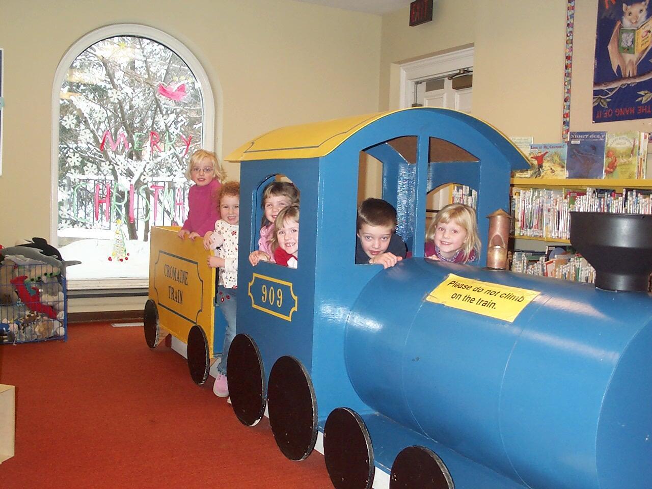 6 young children sit inside a blue and yellow wooden train in the Library 