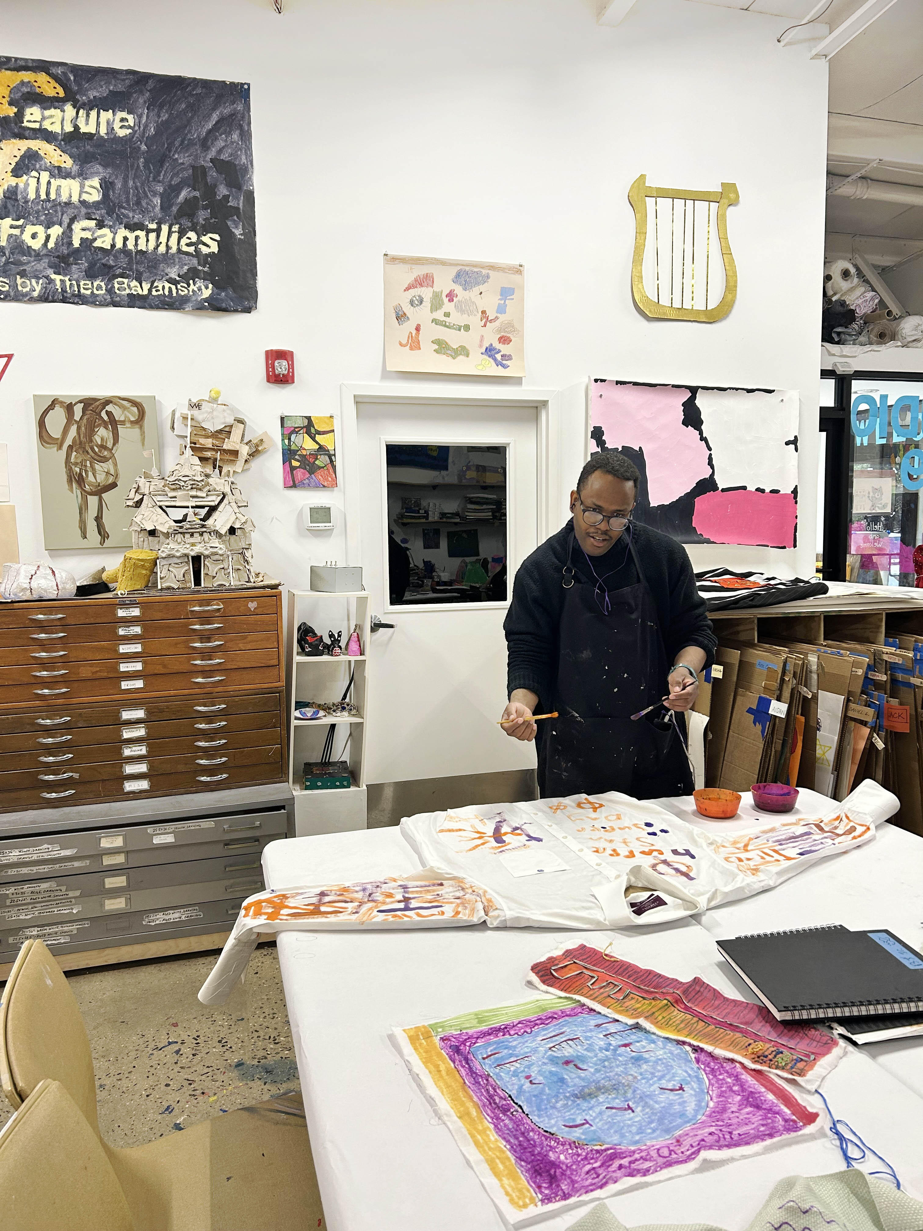 a black man wearing black and an apron holds a brush in each hand while working on a white shirt laid flat on a table in a bright room with art on the walls