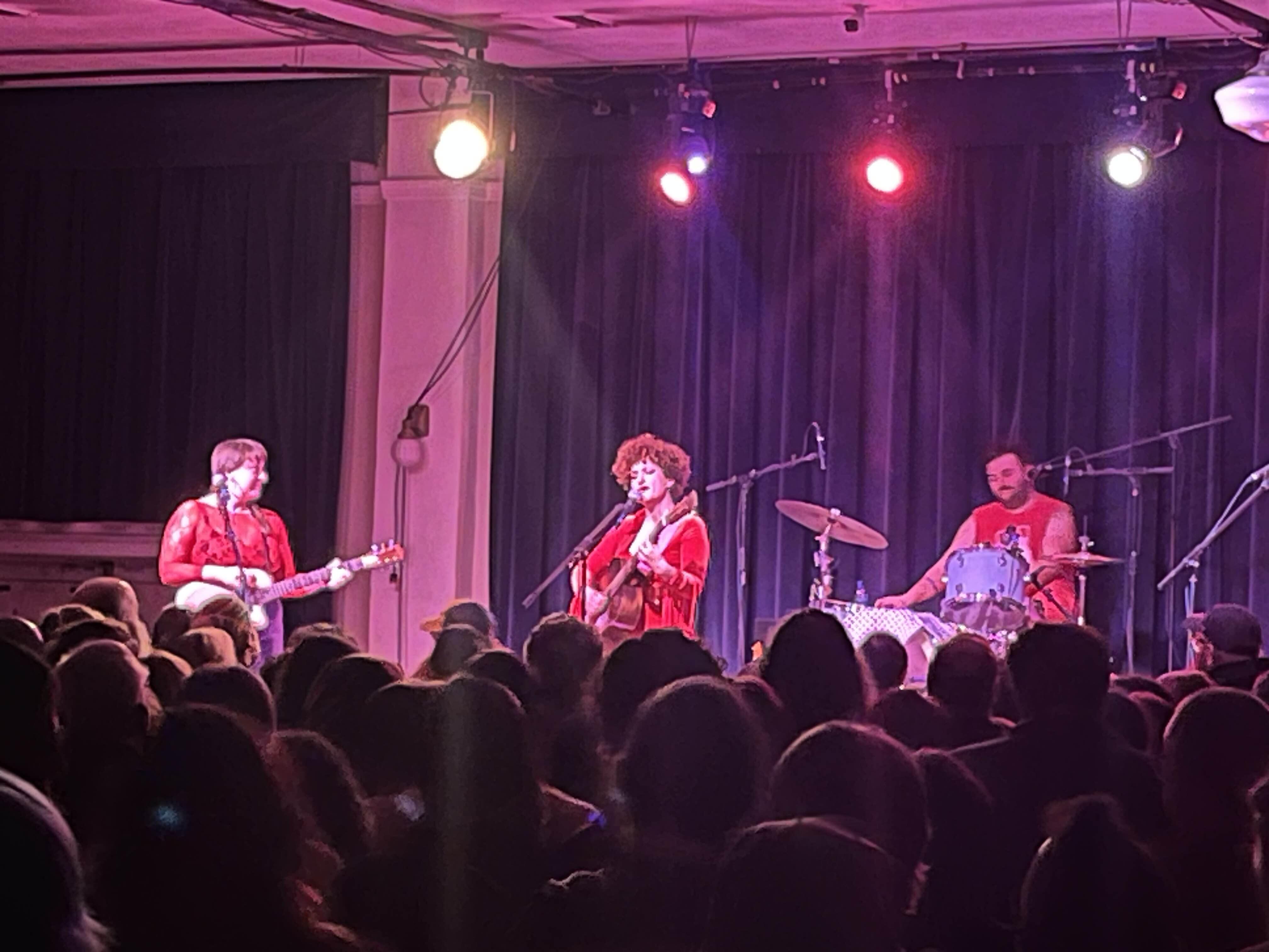 A three-piece band performs on a small indoor stage to a standing-room crowd. Center stage, a woman with curly hair sings into a microphone while playing ukulele, wearing a red velvet top. To her left, a person plays electric guitar; to her right, a mustached drummer sits behind a drum kit. All three musicians wear red. The intimate venue is bathed in warm pink and red stage lighting, with dark curtains as a backdrop and several stage lights visible overhead.